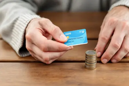 ruka starije osobe drži iskaznicu dopunskog osiguranja pokraj hrpe kovanica što prikazuje cijena dopunskog osiguranja hzzo-a