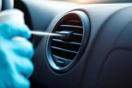 ruka u rukavici čisti ventilacijski otvor u automobilu sprejem prikazujući kako očistiti klimu od bakterija